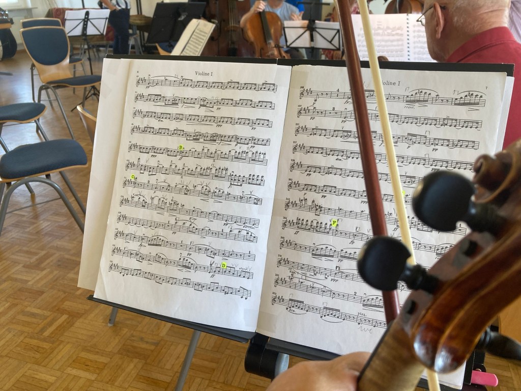 Blick auf ein Notenpult mit Noten für Violine 1, im Vordergrund außerdem die Schnecke meiner Geige und mein Geigenbogen im Bild.